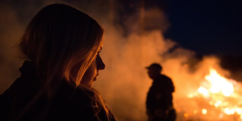 Closeup of woman in foreground and silhouette of man in background with a roaring fire and smoke in the background at night