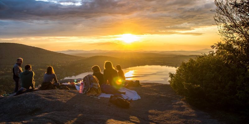 A group of people sitting on a mountain overlooking a lake at sunset