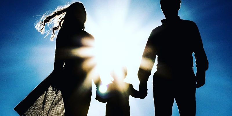 Silhouette of a man and woman holding hands with a child with the sun shinning brightly on them