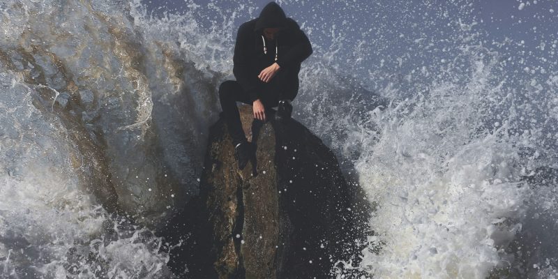 Man in a hoodie sitting on a large rock with is head bowed down being hit with a large ocean wave