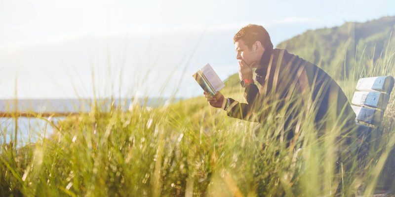 Man sitting outside on a bench near a lake reading the Bible