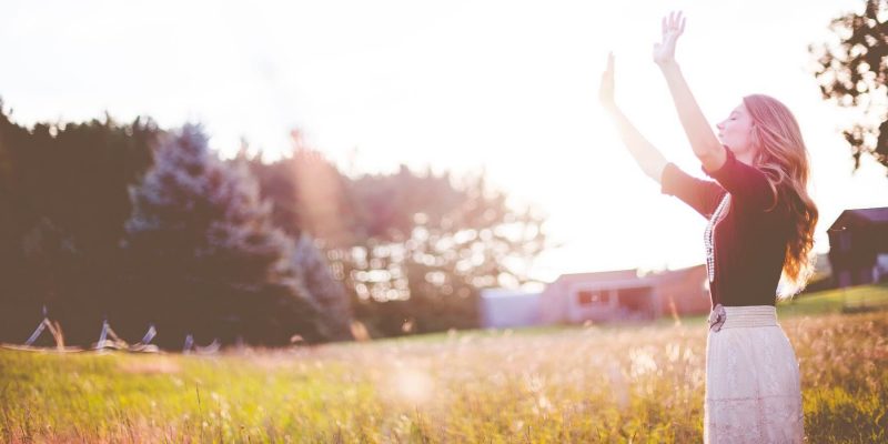 Woman standing in a field with the bright sunshine on her, hands raised up and looking to the sky