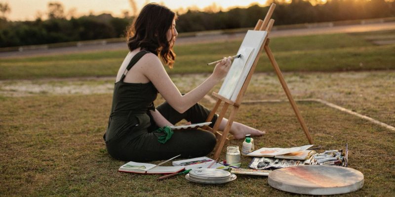 A woman sitting outside on the ground painting on a canvas on an easel.
