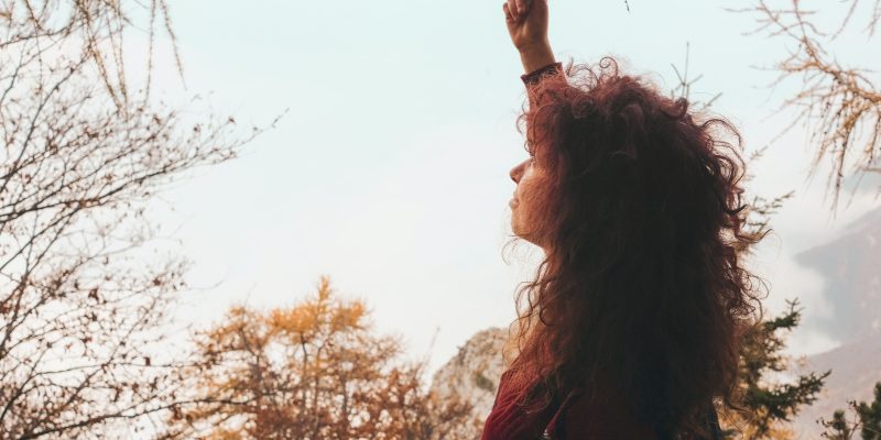 Woman outside looking up to the sky reaching up to hold a tree branch