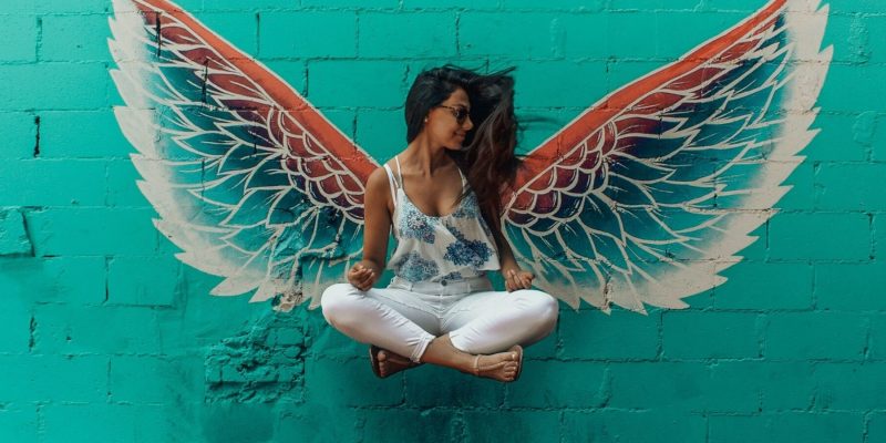 Woman in seated position levitating in the air with angel wings painted on the wall behind her