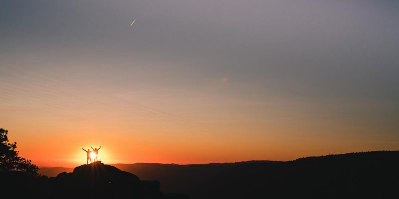 Genuflect - Couple standing on top of a mountain in the distance with sunrise shinning on them