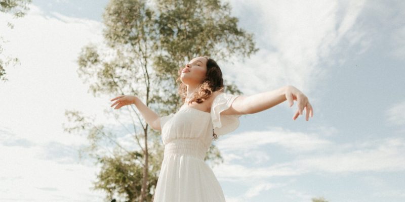 Girl outside on a beautiful day with her face looking up to heaven and eyes closed, arms outstretched as if she is dancing