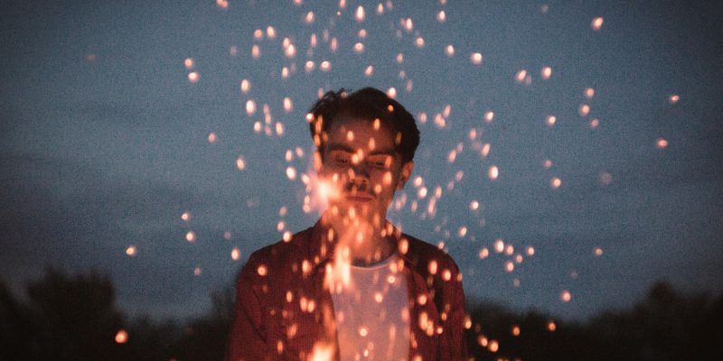 A young man outside at night with sparks from a flame flying up