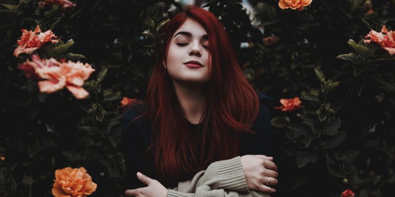 Woman standing outside in front of rose bushes with her eyes closed, arms folded, and a soft smile on her face