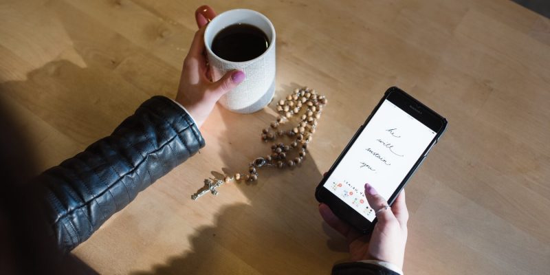 Close up of a table top with a woman holding a cup of coffe and a phone with a rosary sitting on the table