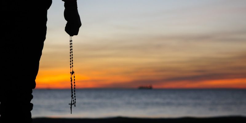 sihouette of a man holding a rosary standing on the beach in front of the sunset over the ocean