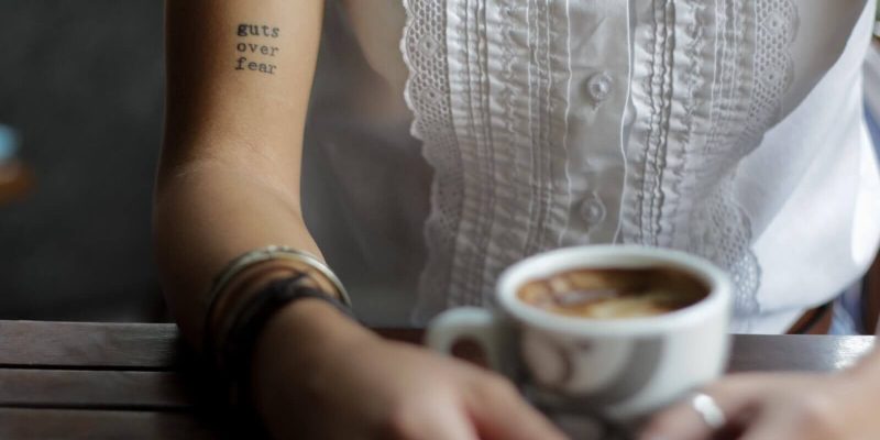 Woman holding a cup of coffee with a tattoo on her arm that reads "guts over fear"