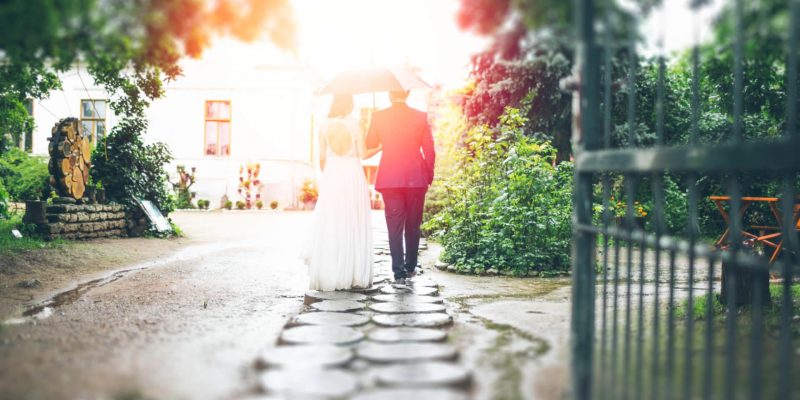 Bride and groom walking outside on a path under an umbrella with the sun shinning brightly on them