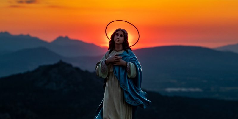 Statue of the Blessed Virgin Mary with a mountain landscape at sunset with an orange sky