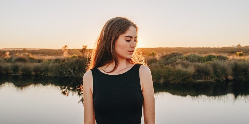 Woman outside at the golden hour standing in front of a body of water looking down contemplatively