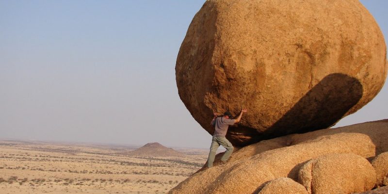A man poses as if holding up a large boulder