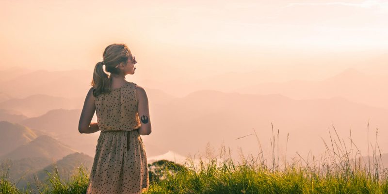 Woman standing in tall grass overlooking the land with a beautiful hazy orange sky