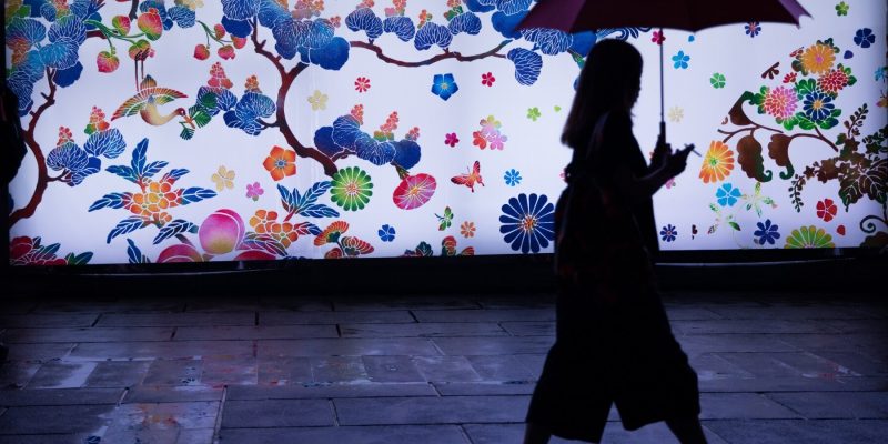 Silhouette of a woman walking with an umbrella in front of a colorful wall with a floral mural
